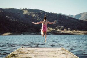 woman wearing pink top jumping towards water during daytime