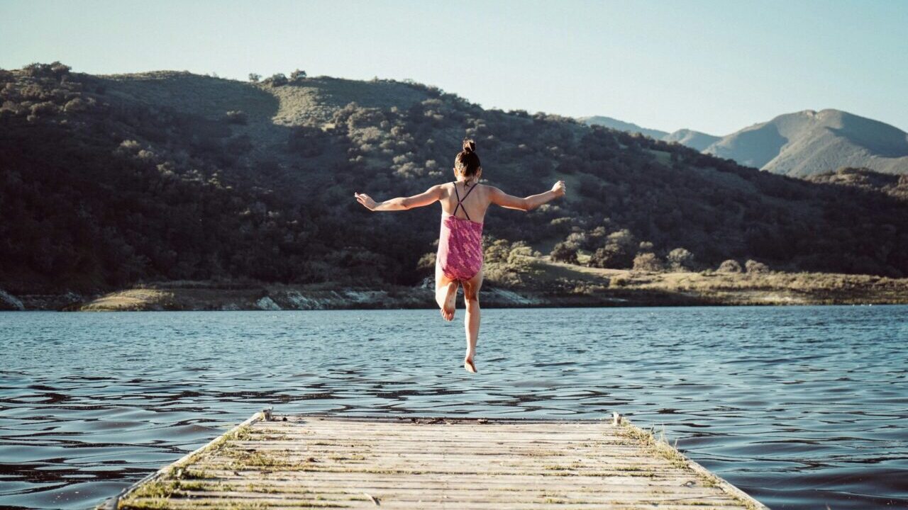 woman wearing pink top jumping towards water during daytime