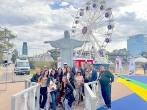 Vibrante grupo de viajantes na feira de turismo no Espaço das Américas, com a famosa estátua do Cristo Redentor e roda gigante ao fundo, promovendo experiências de viagens e turismo no Brasil.