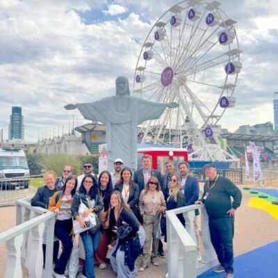 Vibrante grupo de viajantes na feira de turismo no Espaço das Américas, com a famosa estátua do Cristo Redentor e roda gigante ao fundo, promovendo experiências de viagens e turismo no Brasil.