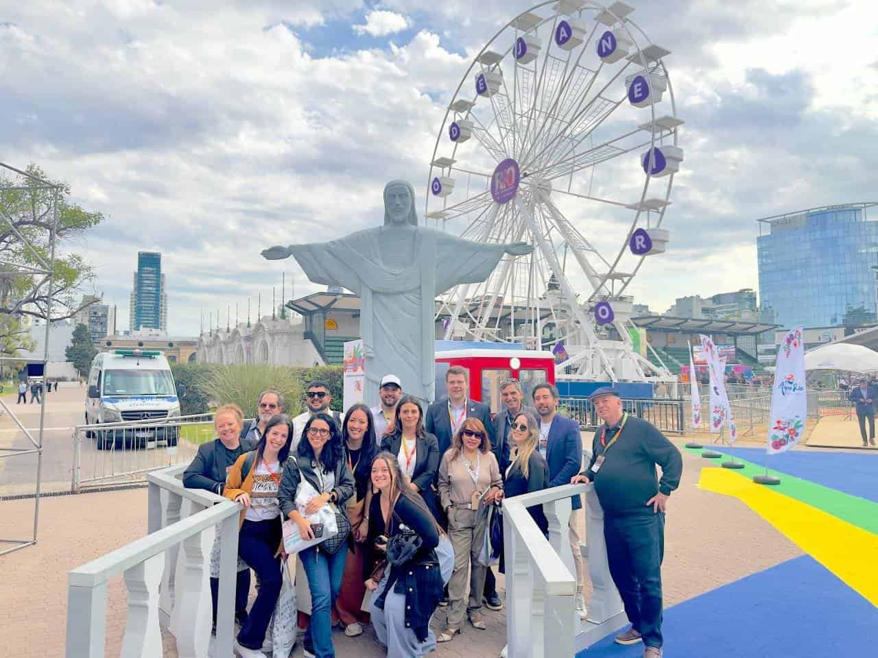 Vibrante grupo de viajantes na feira de turismo no Espaço das Américas, com a famosa estátua do Cristo Redentor e roda gigante ao fundo, promovendo experiências de viagens e turismo no Brasil.