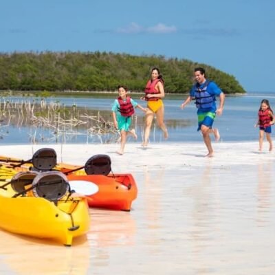 Pai, mãe e duas crianças com coletes salva-vidas na praia, desembarcando de caiaques coloridos em águas rasas, com ilha e céu azul ao fundo, promovendo turismo de aventura e lazer na natureza.