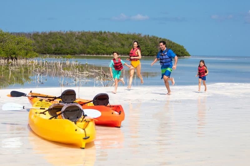 Pai, mãe e duas crianças com coletes salva-vidas na praia, desembarcando de caiaques coloridos em águas rasas, com ilha e céu azul ao fundo, promovendo turismo de aventura e lazer na natureza.