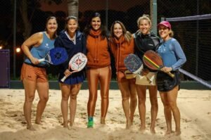 Equipe de natação feminina posando após treino ou competição com rackets de beach tennis em quadra de areia, destacando esportes de praia, saúde e bem-estar, em ambiente ao ar livre.