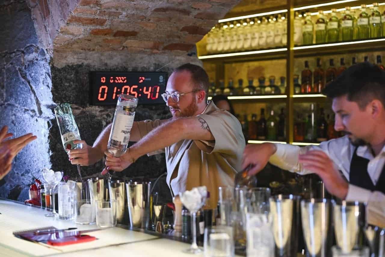 Primeiro plano de bartenders preparando drinks em bar moderno com iluminação aconchegante e decoração em tijolos expostos, promovendo experiências de entretenimento e gastronomia sofisticada.