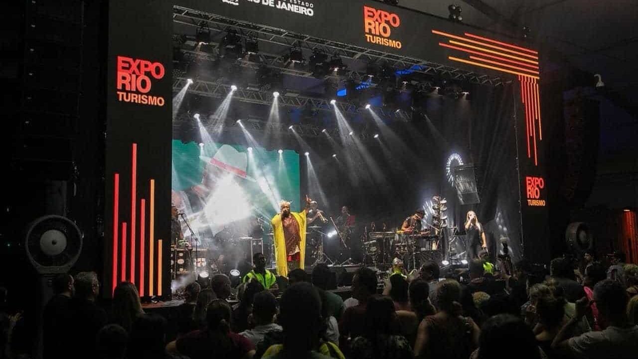 Público assistindo a show no evento de turismo Expo Rio Rio Janeiro, com palco iluminado, banda performing e destaque para o cenário de feira de turismo da cidade.