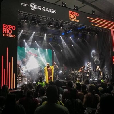 Público assistindo a show no evento de turismo Expo Rio Rio Janeiro, com palco iluminado, banda performing e destaque para o cenário de feira de turismo da cidade.