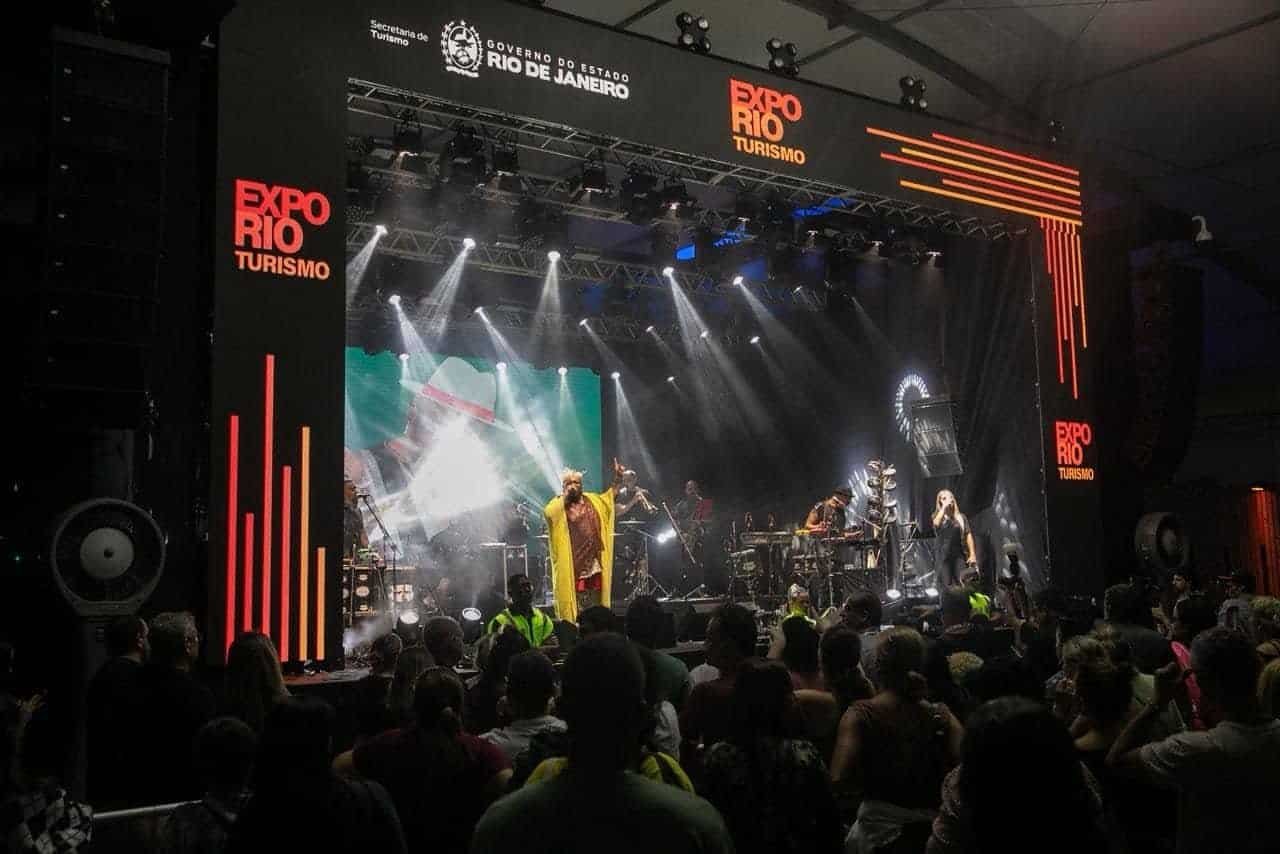 SuoViaggio - Evento de turismo no Rio de Janeiro com show ao vivo, iluminação profissional e grande público apreciando música e cultura local em uma feira de turismo e experiências culturais. Público assistindo a show no evento de turismo Expo Rio Rio Janeiro, com palco iluminado, banda performing e destaque para o cenário de feira de turismo da cidade.